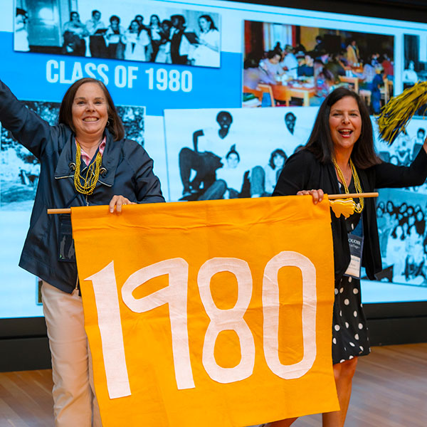 The class of 1980 students hold a celebratory yellow banner reading "1980" while waving pom poms at the '25 Alumni Weekend.