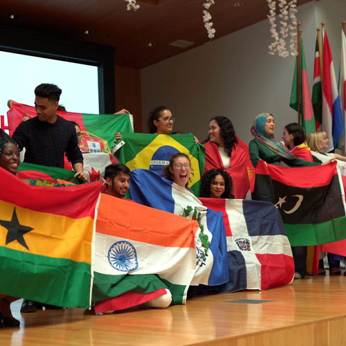 A group of international students, standing on the Hyman Forum stage, excitedly holding flags of their home countries during the 2024 International Festival.